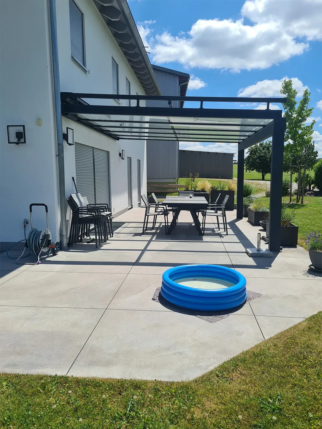 offene Terrasse mit Glasüberdachung, Gartenmöbeln und blauem Kinderplanschbecken