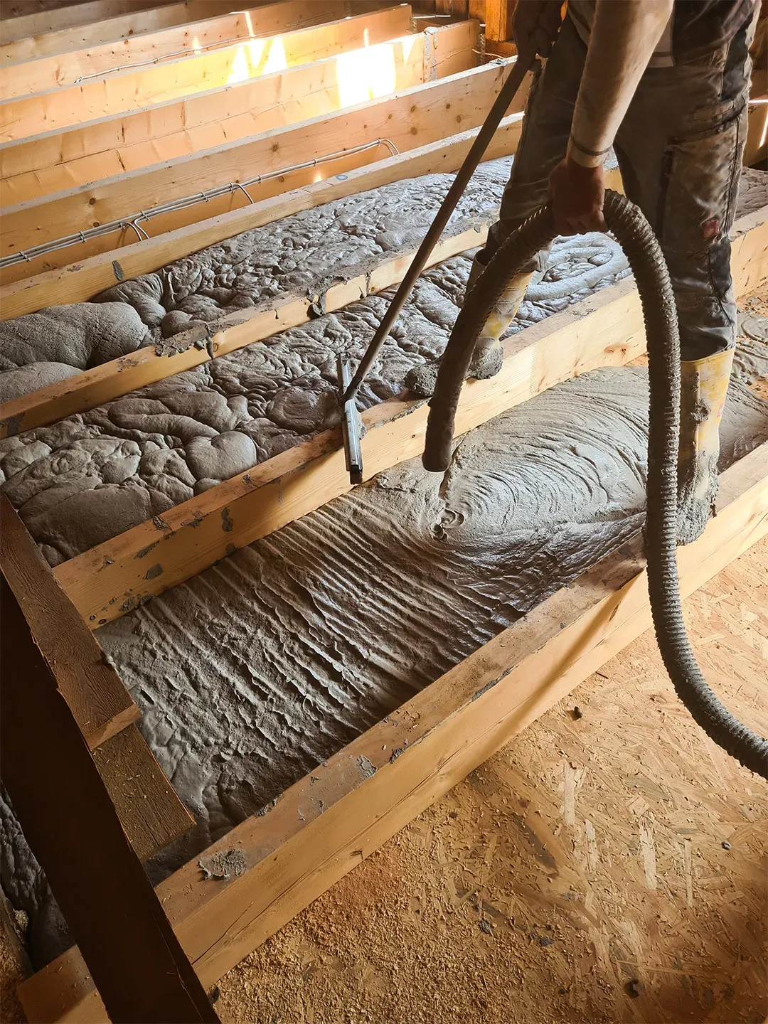 Ein Bodenaufbau mit sichtbaren Holzbalken, die mit frischer Schüttdämmung befüllt werden, ein Arbeiter mit einem Schlauchwerkzeug im Einsatz