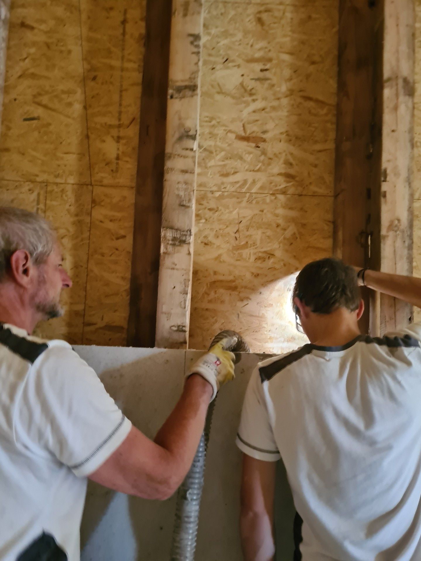 Zwei Personen arbeiten an einer Wandkonstruktion, einer führt einen flexiblen Schlauch in einen Wandhohlraum ein