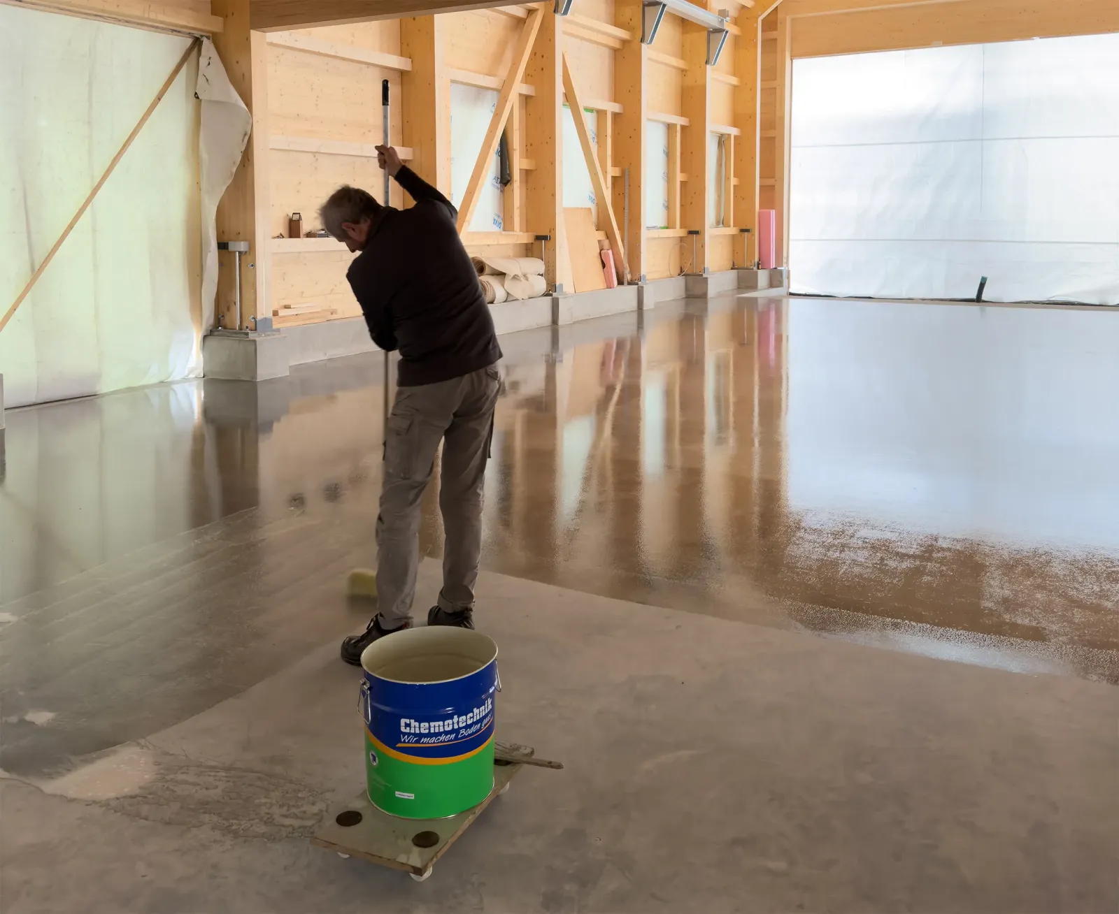Rohbauhalle mit Holzwänden, ein Arbeiter verteilt Flüssigkeit mit einer Rolle auf einem frisch beschichteten Boden