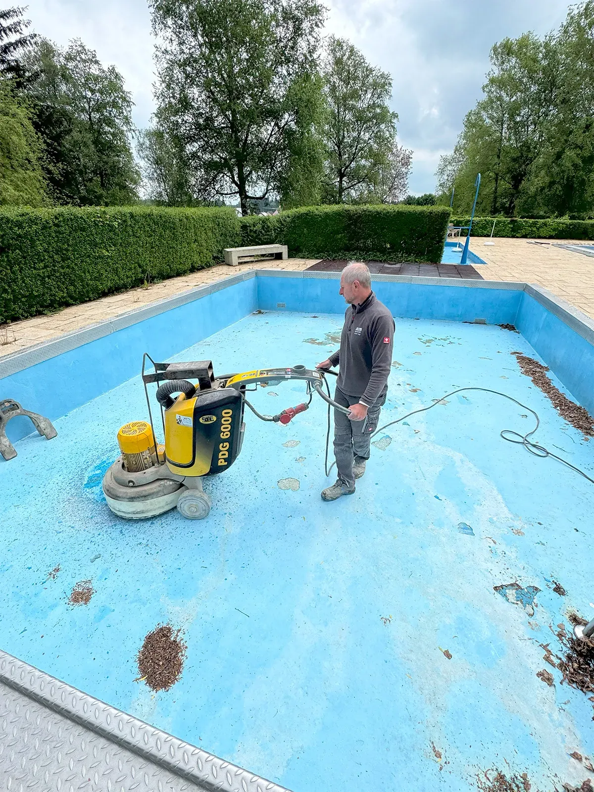 Arbeiter verwendet Schleifmaschine zur Bearbeitung eines leeren, blauen Schwimmbeckens in einer Außenanlage