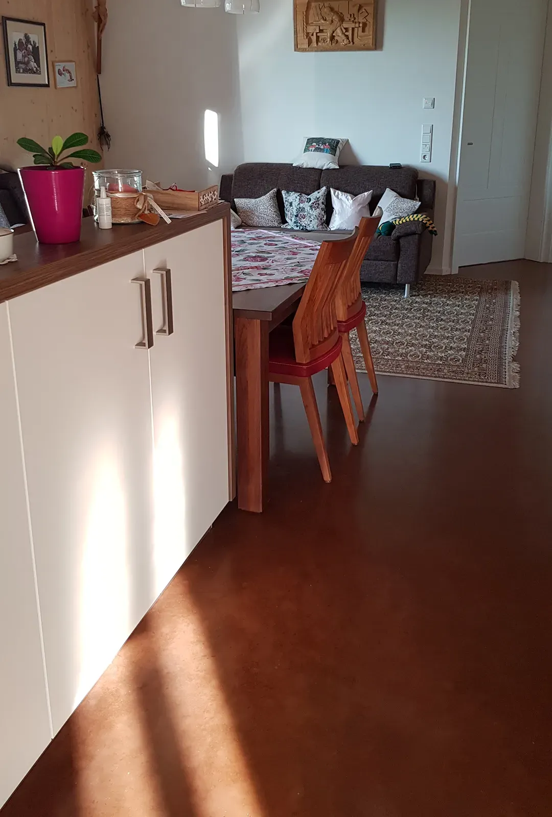 Ein Innenraum mit einem warmen Holzboden, Küchenschränken, einem Esstisch mit roten Stühlen und einer Couch im Hintergrund