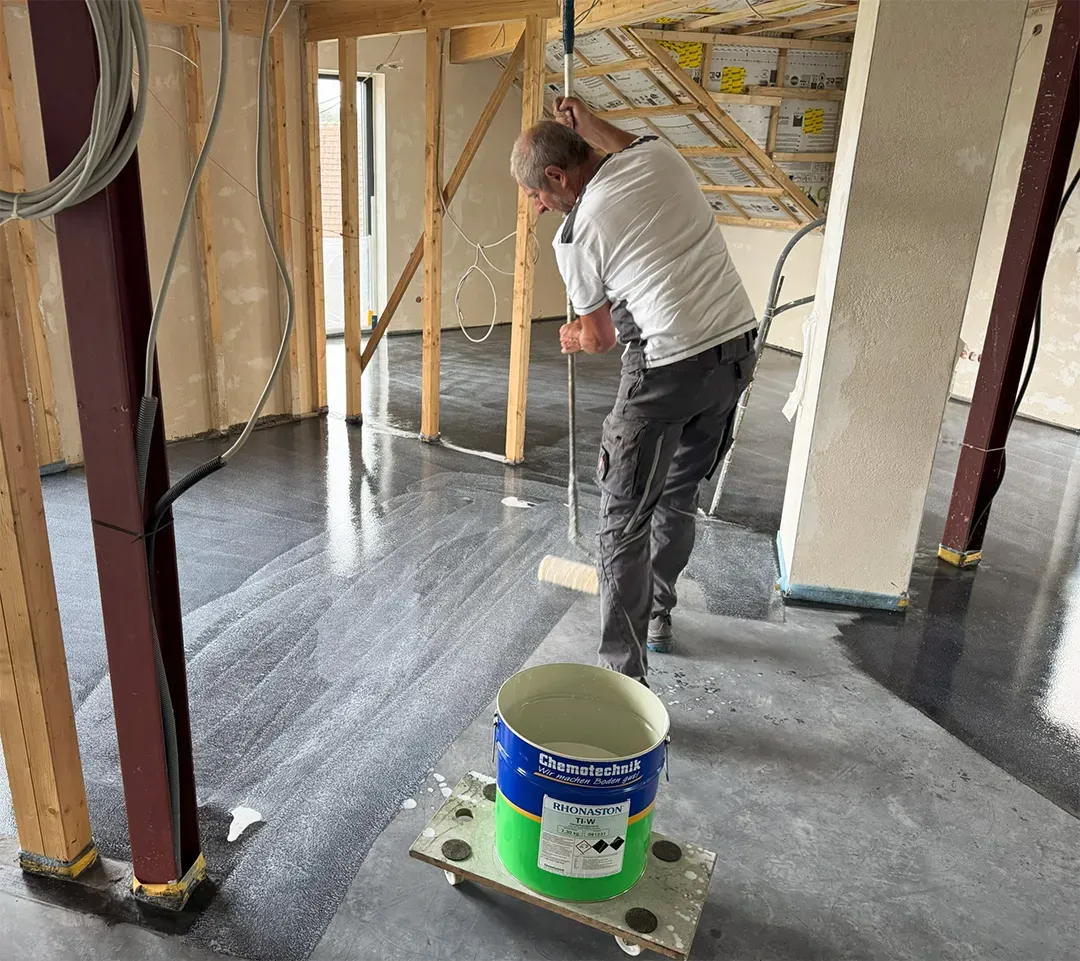 Ein Rohbau mit Holzbalken und frei sichtbarer Decke, ein Arbeiter verteilt eine schwarze Flüssigkeit mit einer Rolle auf dem Boden