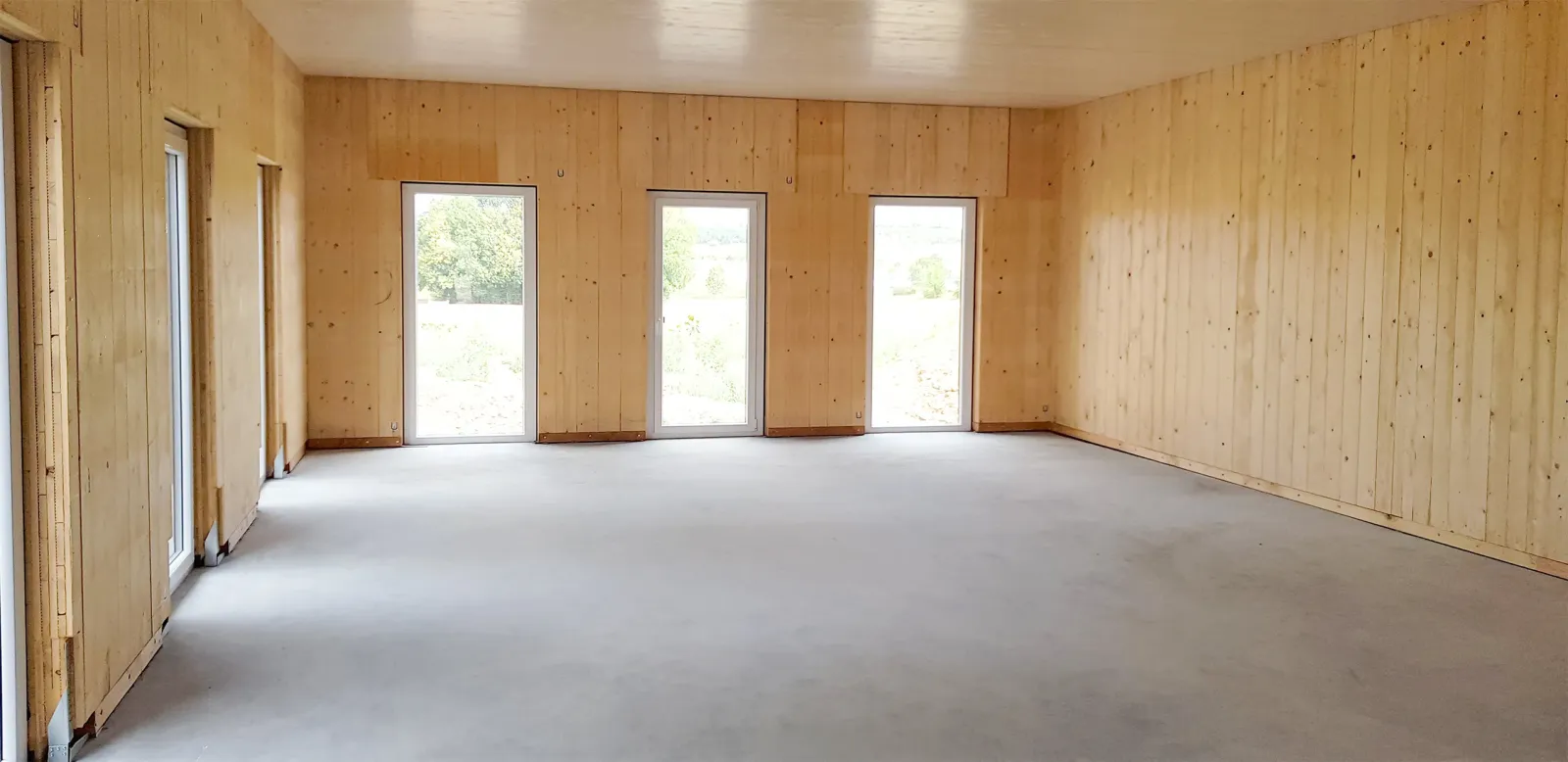 Ein leerer Raum mit Holzverkleidung an Wänden und Decke, großen bodentiefen Fenstern und einem glatten Betonboden