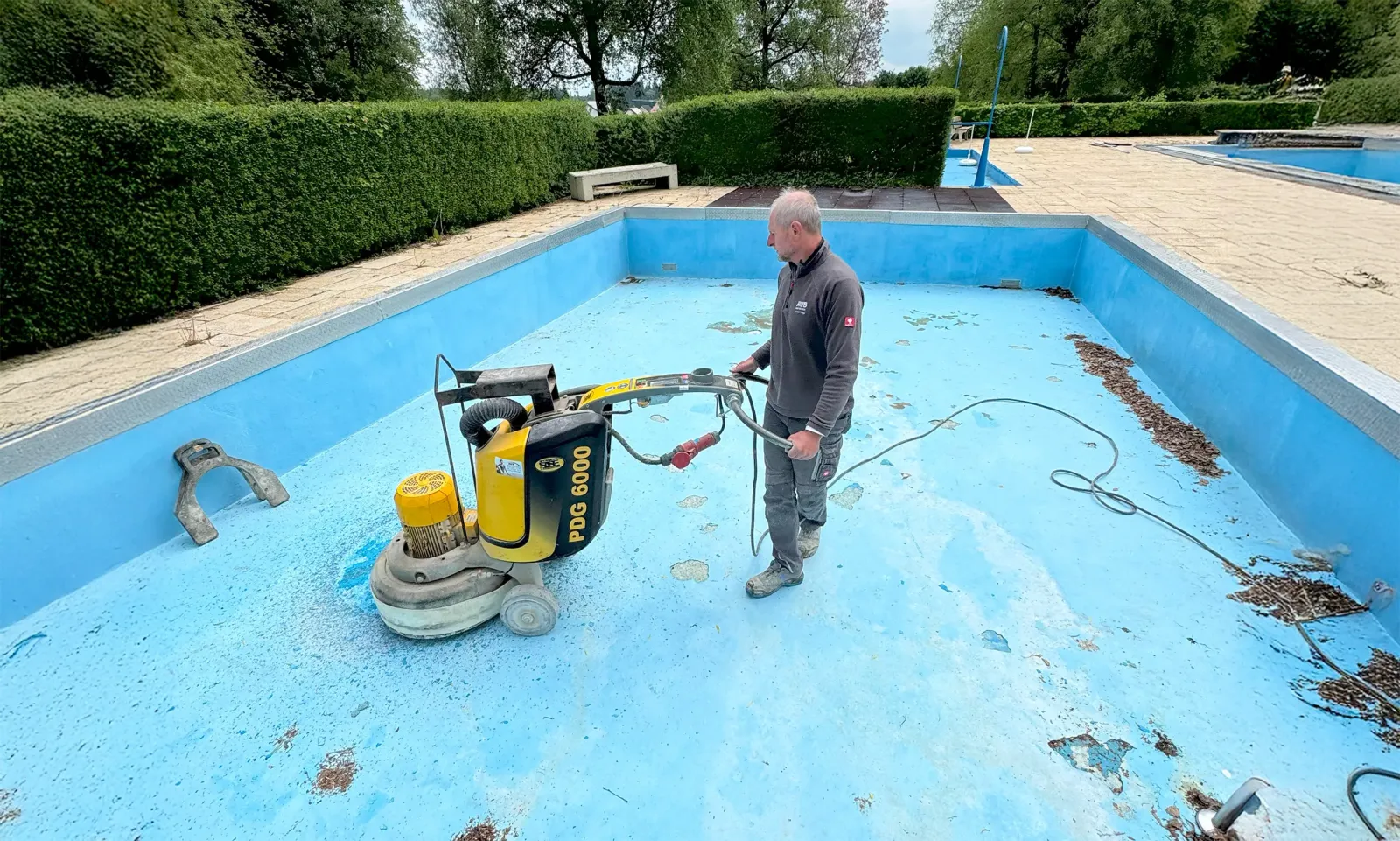 Leeres Schwimmbecken mit blauen Wänden, ein Arbeiter bedient eine Schleifmaschine zur Bodenbearbeitung