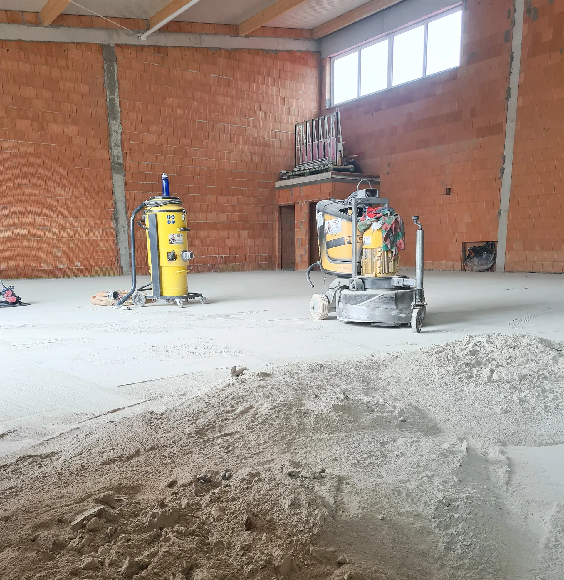 Betonboden in Bauphase mit Schleifmaschinen und gelbem Staubsauger in einer Halle mit unverputzten Wänden