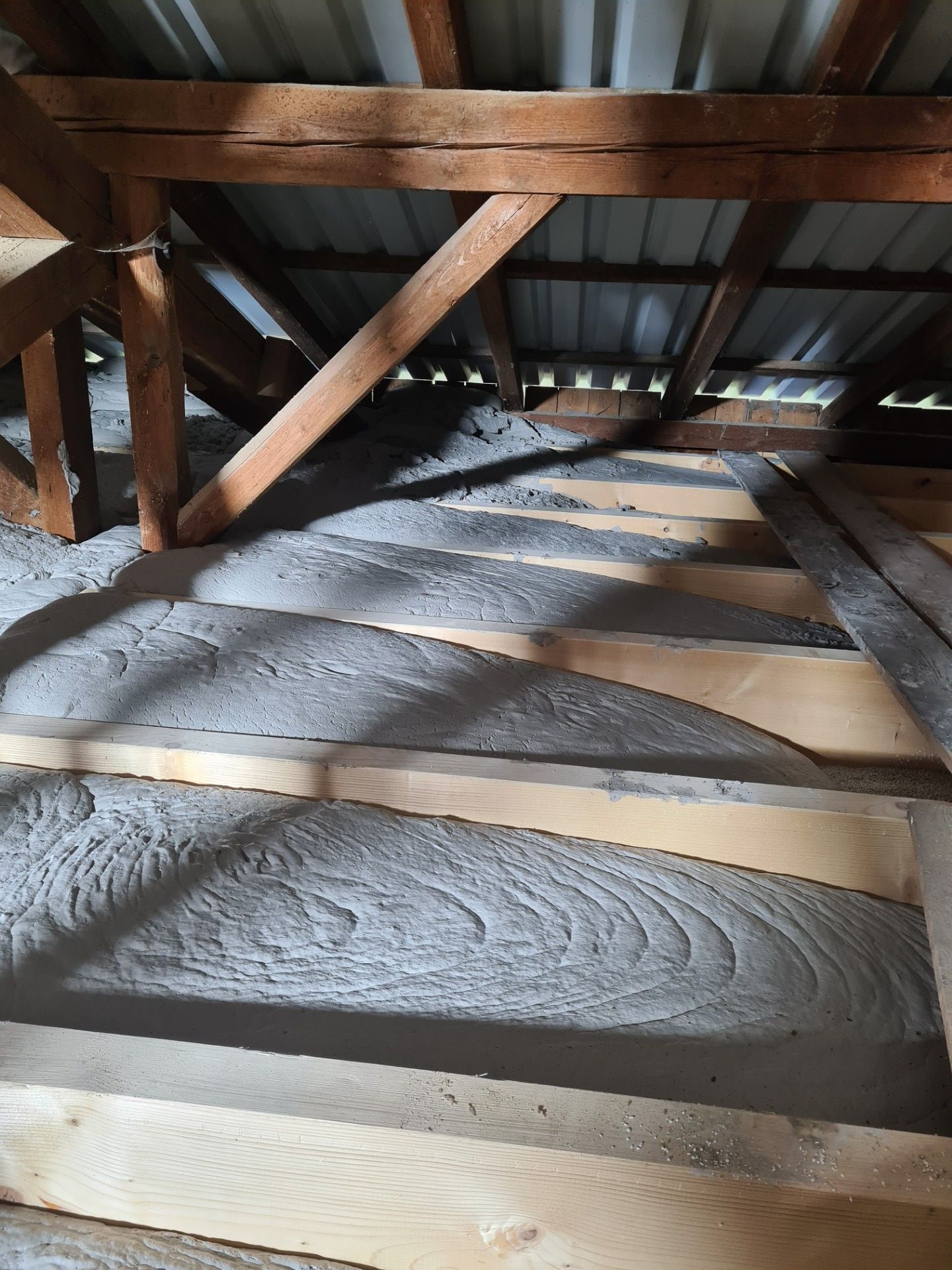 Ein Dachboden mit Holzbalken, die mit frischer Schüttdämmung befüllt werden, ein Arbeiter mit einem Werkzeug in Aktion